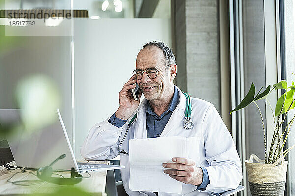 Lächelnder reifer Arzt mit medizinischem Bericht  der am Schreibtisch auf dem Smartphone spricht