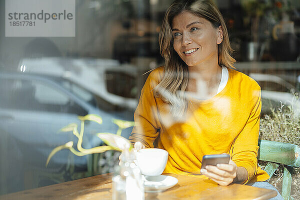 Glückliche Frau beim Kaffeetrinken im Café  gesehen durch Glas