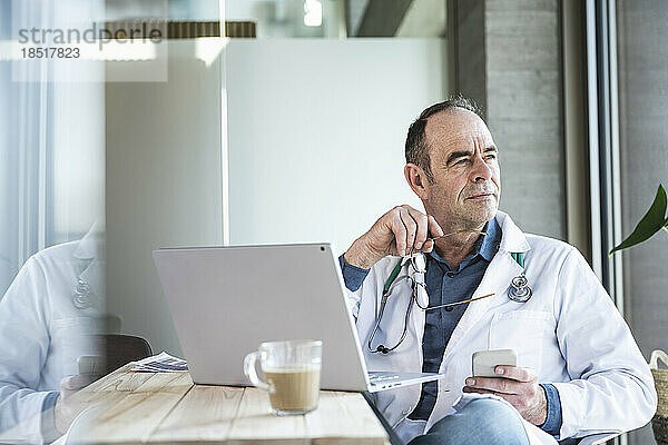 Nachdenklicher Arzt mit Brille und Mobiltelefon sitzt am Schreibtisch