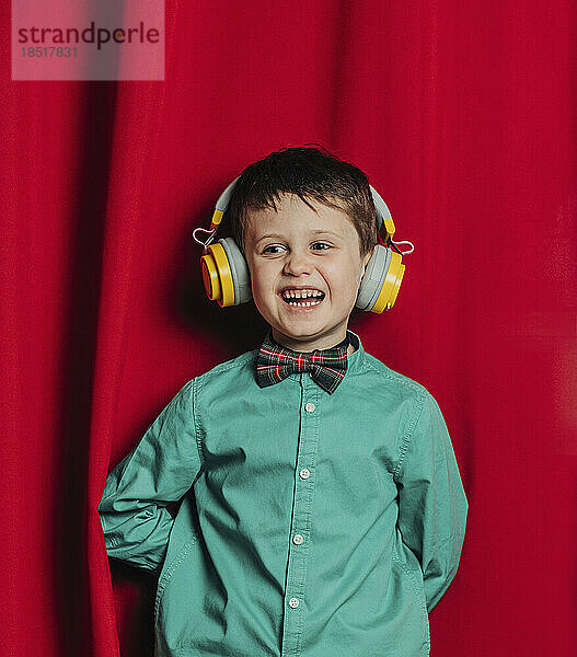 Fröhlicher Junge mit kabellosen Kopfhörern steht vor rotem Vorhang
