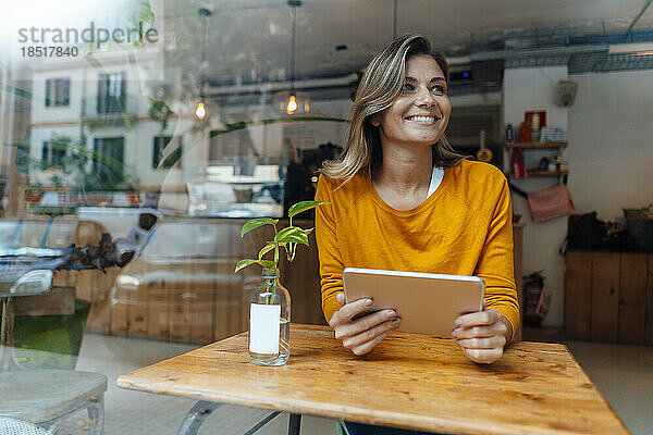 Glückliche Frau sitzt mit Tablet-Computer im Café und sieht durch Glas