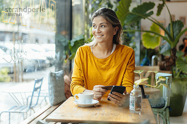 Glückliche Frau mit Kreditkarte und Mobiltelefon sitzt im Café
