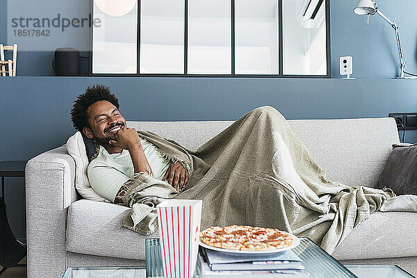 Lächelnder Mann mit Decke entspannt sich zu Hause auf dem Sofa