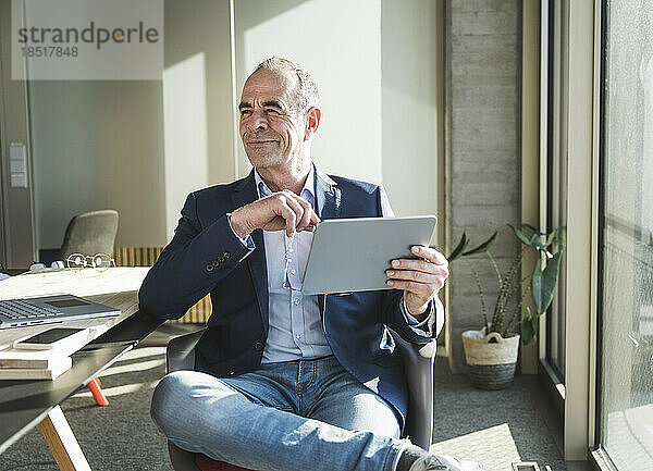 Nachdenklich lächelnder Geschäftsmann mit Tablet-PC sitzt im Büro
