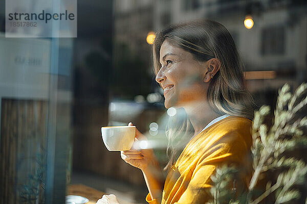 Nachdenklich lächelnde Frau beim Kaffeetrinken im Café  gesehen durch Glas