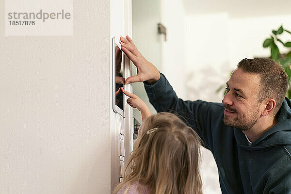 Lächelnder Vater blickt seine Tochter an  die zu Hause die Hausautomations-App auf einem Tablet-PC nutzt