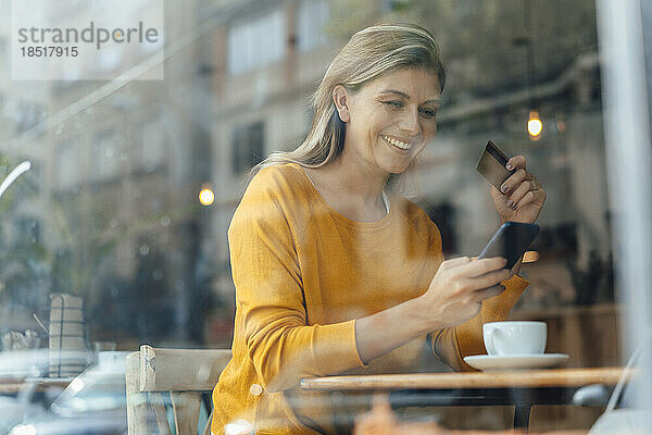 Glückliche Frau benutzt Smartphone mit Kreditkarte im Café  gesehen durch Glas