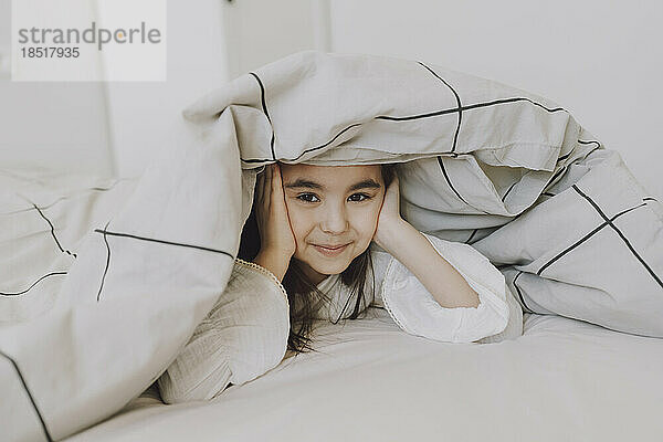 Smiling cute girl lying under blanket on bed