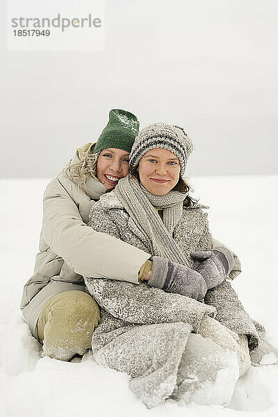 Glückliche Frau umarmt Freundin  die im Winter im Schnee sitzt