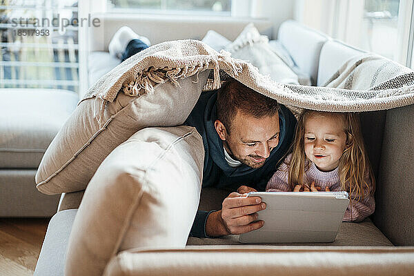Glücklicher Vater und Tochter schauen auf den Tablet-PC unter einer Decke  die zu Hause auf dem Sofa liegt