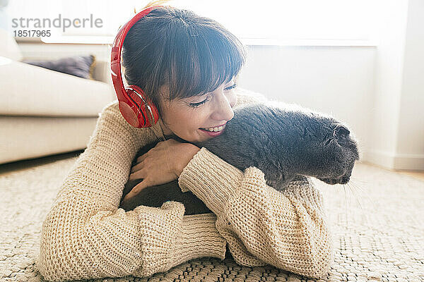 Glückliche Frau im Pullover umarmt die Katze  die zu Hause auf dem Teppich liegt