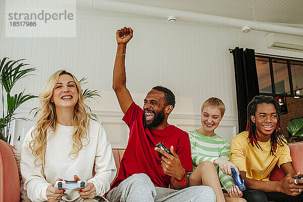 Aufgeregter junger Mann mit erhobener Hand sitzt neben Freunden und spielt zu Hause ein Videospiel