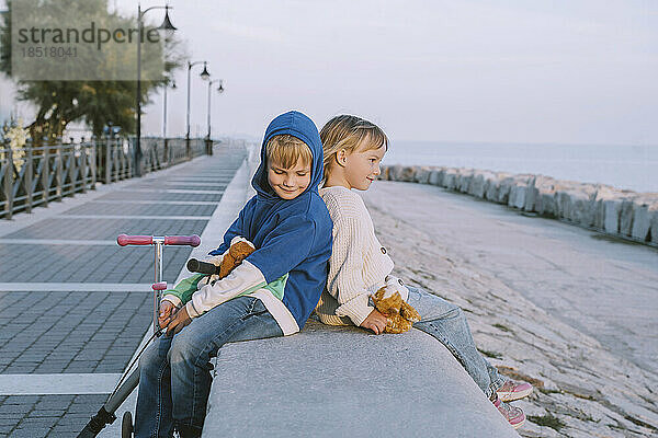 Lächelnder Junge sitzt mit Schwester auf der Promenade bei Sonnenuntergang