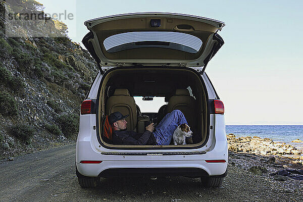 Mann benutzt Smartphone im Urlaub im Kofferraum eines Autos