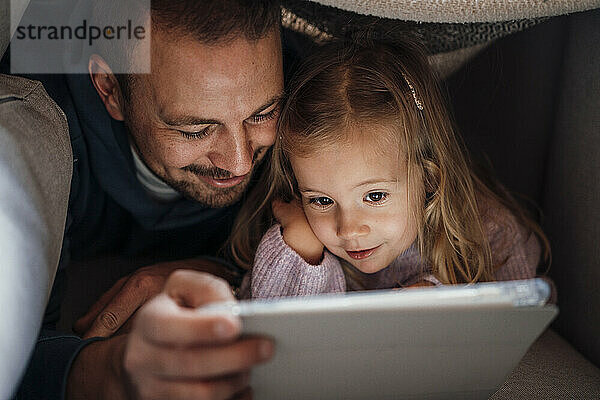 Vater und Tochter schauen zu Hause unter der Decke auf den Tablet-PC