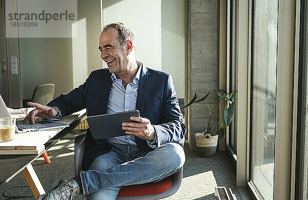 Glücklicher Geschäftsmann benutzt Laptop und hält Tablet-PC im Büro