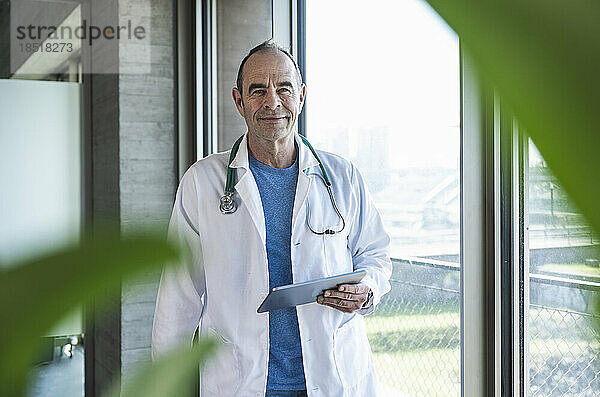 Lächelnder reifer Arzt steht mit Tablet-PC am Fenster