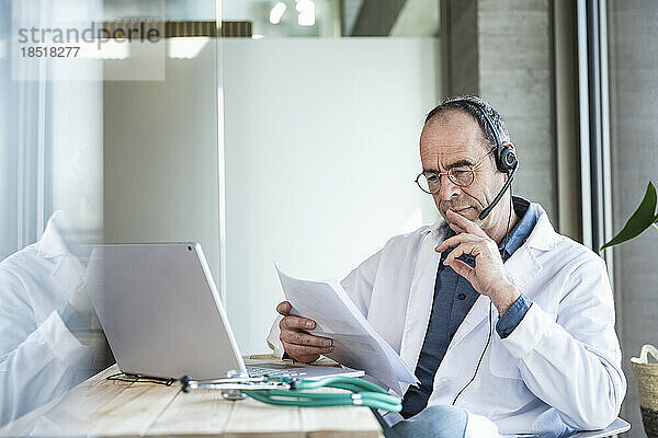 Arzt mit Headset liest Dokument und sitzt mit Laptop im Büro