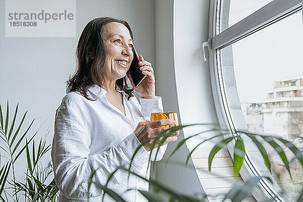 Glückliche Geschäftsfrau spricht mit Smartphone und hält Glas am Fenster
