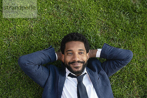 Thoughtful smiling businessman with hands behind head lying on grass