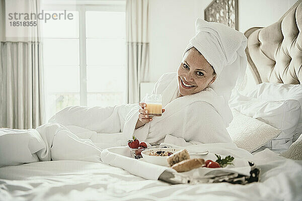 Glückliche Frau hält ein Saftglas neben dem Frühstückstablett auf dem heimischen Bett