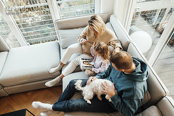 Tochter schaut sich Fotos mit Eltern und Hund an  die zu Hause auf dem Sofa sitzen