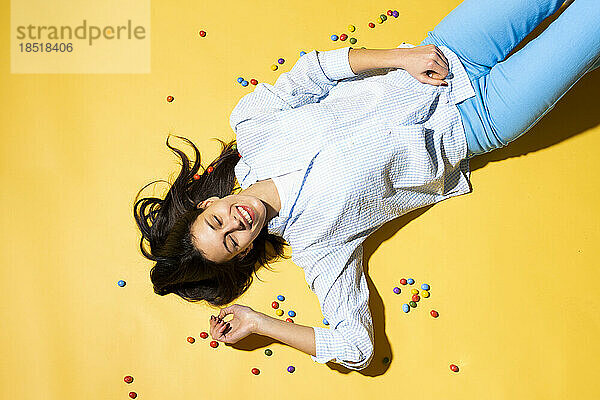 Happy woman lying amidst colorful candies against yellow background