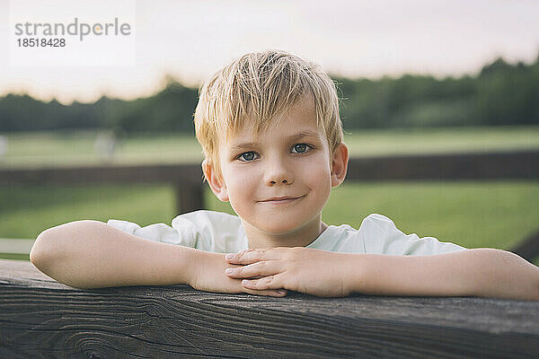Lächelnder blonder Junge lehnt an Holzzaun