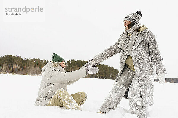 Glückliche Frau zieht Freundin  die im Winter auf Schnee sitzt