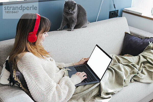 Frau mit kabellosen Kopfhörern schaut zu Hause auf dem Sofa auf den Laptop