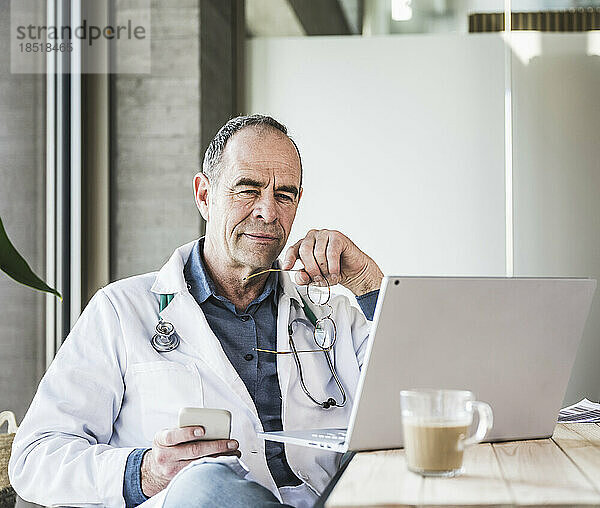 Reifer Arzt hält Smartphone in der Hand und schaut am Laptop zu  der am Schreibtisch sitzt
