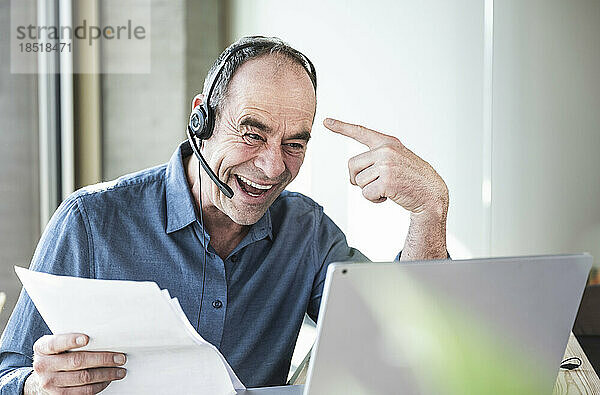 Glücklicher reifer Geschäftsmann mit Headset  der im Büro über Laptop gestikuliert