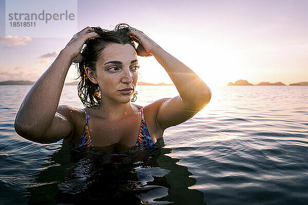 Nachdenkliche junge Frau mit der Hand im Haar im Wasser bei Sonnenuntergang