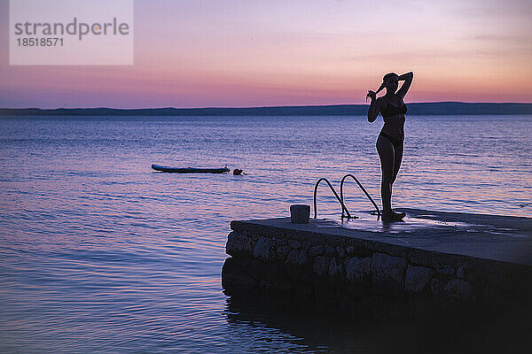 Silhouette einer Frau  die bei Sonnenuntergang auf einem Steg am Meer steht
