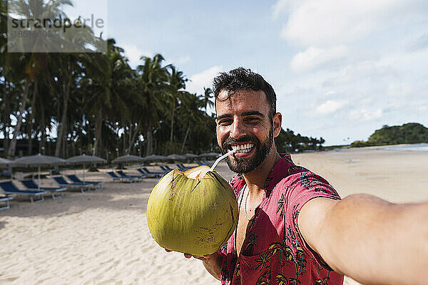 Glücklicher Mann trinkt Kokosnusswasser und macht ein Selfie am Strand