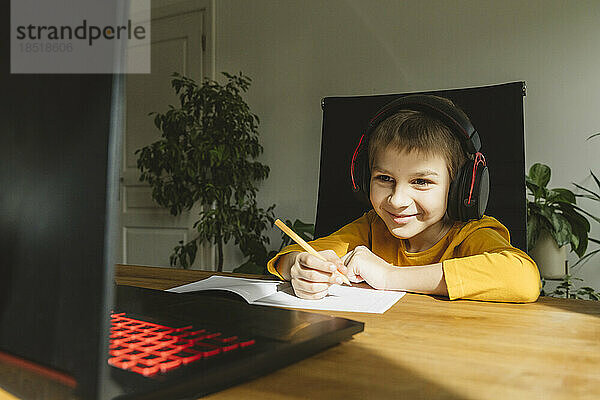 Lächelnder Junge mit Headset  der zu Hause online über den Laptop lernt