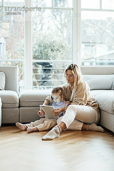 Frau teilt Tablet-PC mit Tochter  die im Wohnzimmer auf dem Boden sitzt