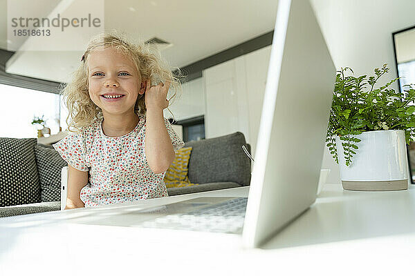 Smiling girl with hand in hair sitting by laptop at home