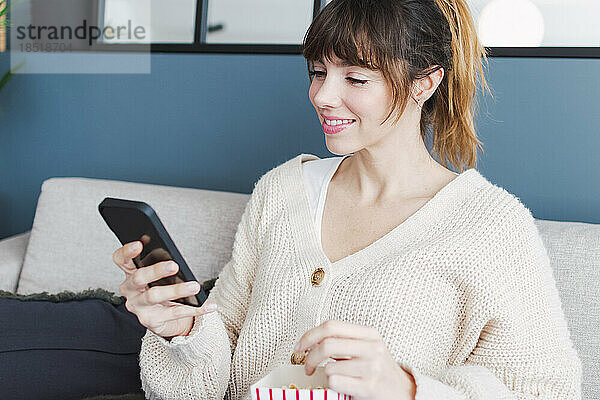Lächelnde Frau  die zu Hause auf dem Sofa sitzt und ihr Smartphone benutzt