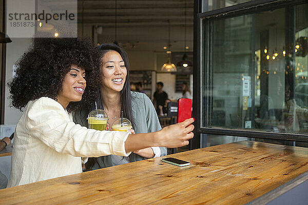Lächelnde Frau macht ein Selfie mit einer Freundin  die im Café ein gesundes Getränk in der Hand hält