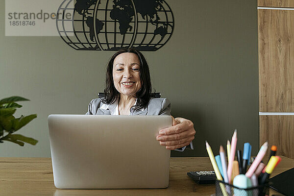 Glückliche Geschäftsfrau öffnet Laptop am Schreibtisch im Büro