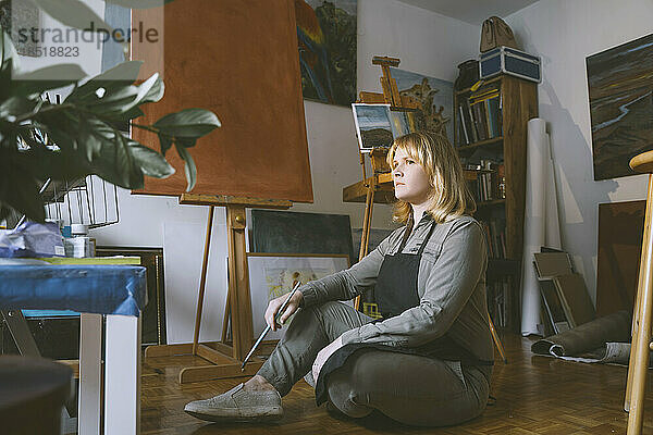 Nachdenklicher Maler mit Pinsel auf dem Boden im Kunstatelier sitzend