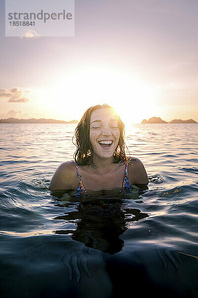 Glückliche Frau mit geschlossenen Augen schwimmt bei Sonnenuntergang im Meer