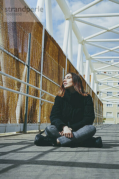 Frau sitzt an einem sonnigen Tag mit gekreuzten Beinen auf einer Brücke