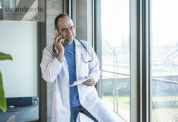 Lächelnder reifer Arzt  der am Fenster steht und am Smartphone spricht