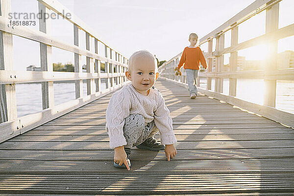 Netter Junge kauert auf einer Holzbrücke  während seine Schwester im Hintergrund läuft