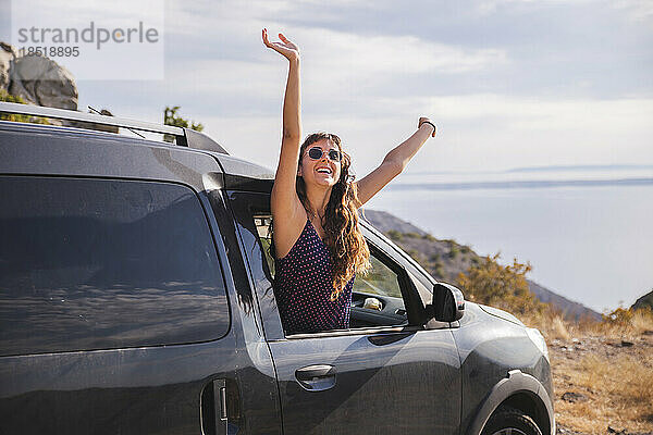 Fröhliche junge Frau lehnt mit erhobenen Armen aus dem Autofenster