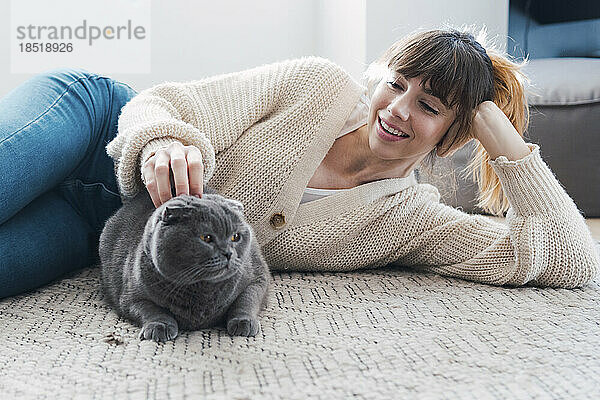Lächelnde Frau streichelt Katze  die zu Hause auf dem Teppich liegt