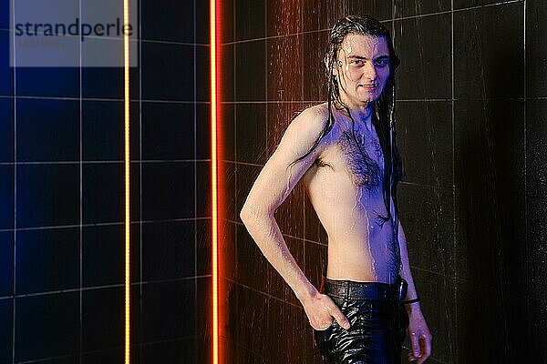 Fröhlicher Mann mit langen Haaren posiert unter Wasserdüsen in Dusche mit Neonlicht