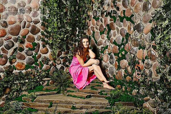 Eine erwachsene Frau sitzt in ihrem hübschen Sommerkleid  mit gewelltem Haar und einem charmanten Lächeln auf einer Steinplatte nahe der Mauer. Sie sieht entspannt und zufrieden aus und genießt das Wochenende im Freien